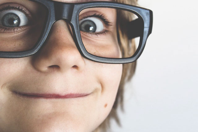 παιδί με γυαλιά που κοιτάει με βλέμμα σκανταλιάς στο φακό