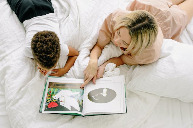 μητέρα με παιδί ξαπλωμένοι μπρούμητα σε κρεβάτι βλέποντας ένα βιβλίο με χρωματιστές εικόνες